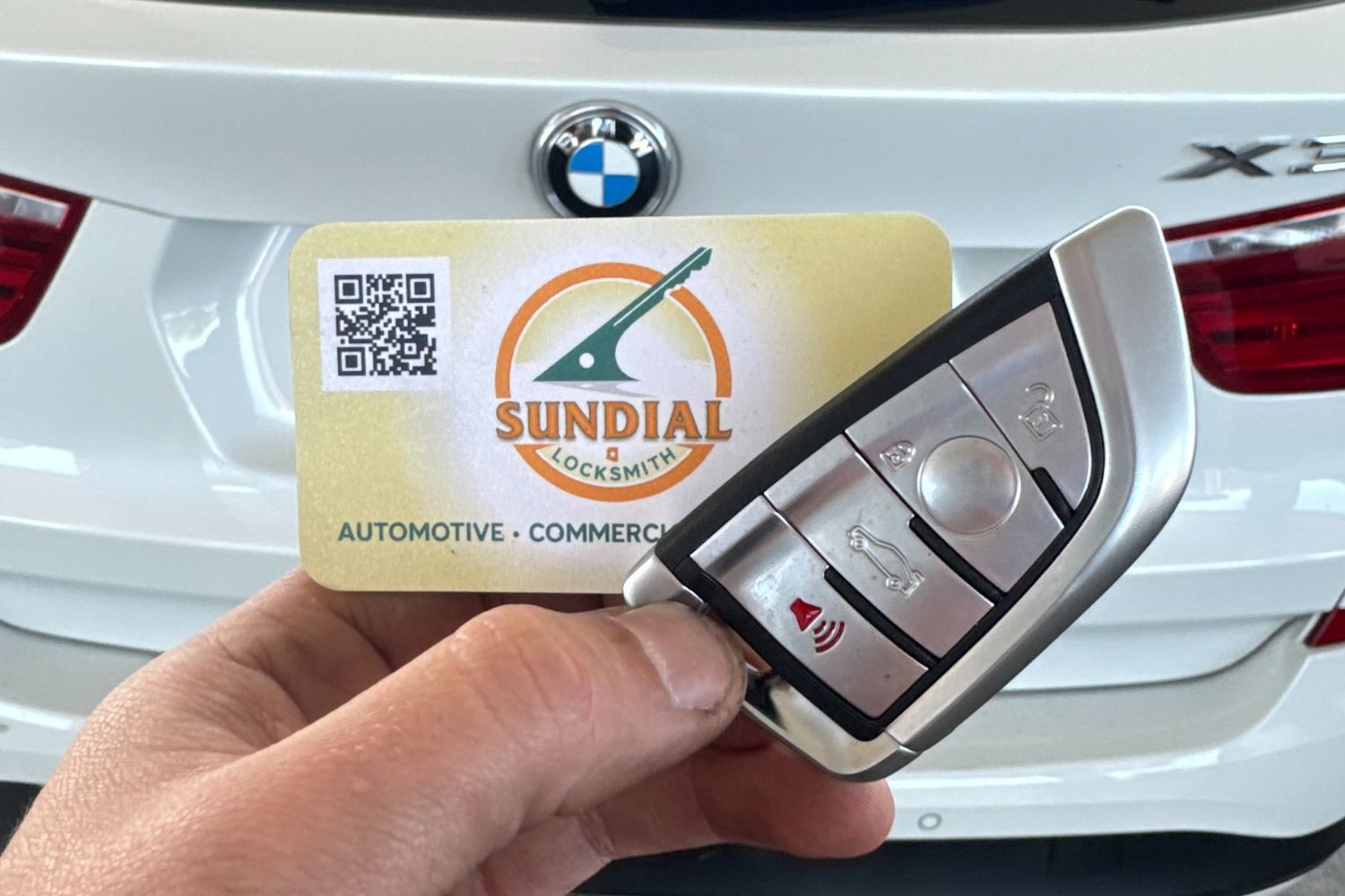 A Sundial Locksmith key fob replacement displayed with a business card in front of a white BMW X3.