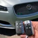 Hand holding two Jaguar key fobs in front of a silver Jaguar vehicle's front grille and headlight, with a 'SUNDIAL' business card placed on the grille