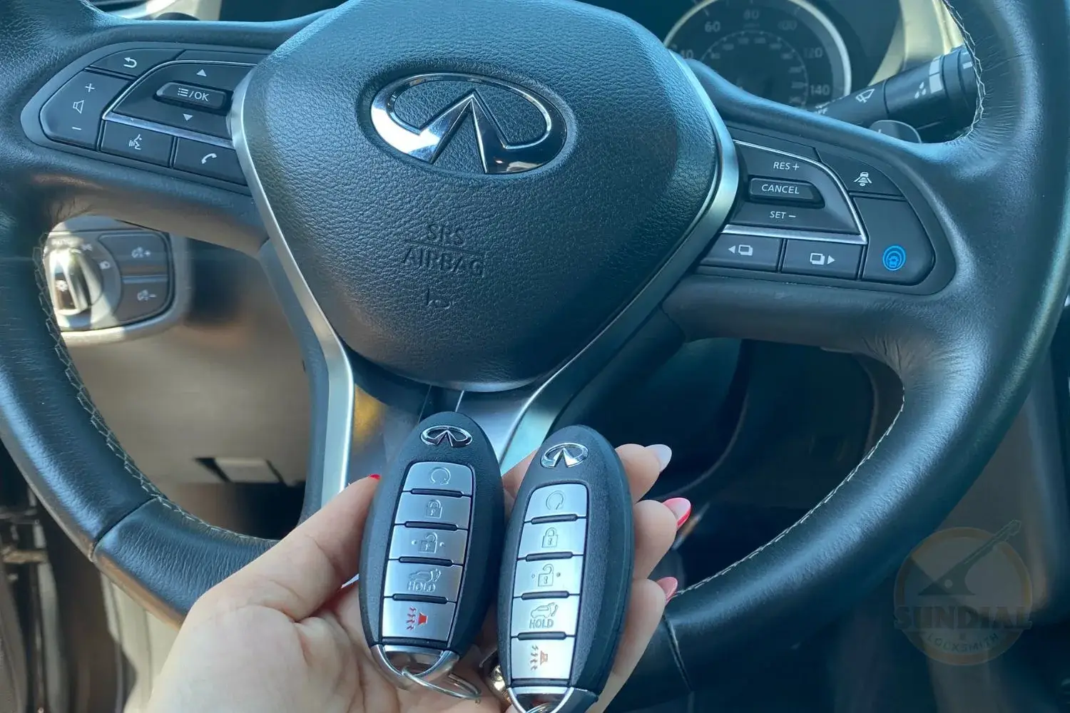 A person's hand showcasing two car key fobs against a luxury car's steering wheel with control buttons and instrument panel in the background.