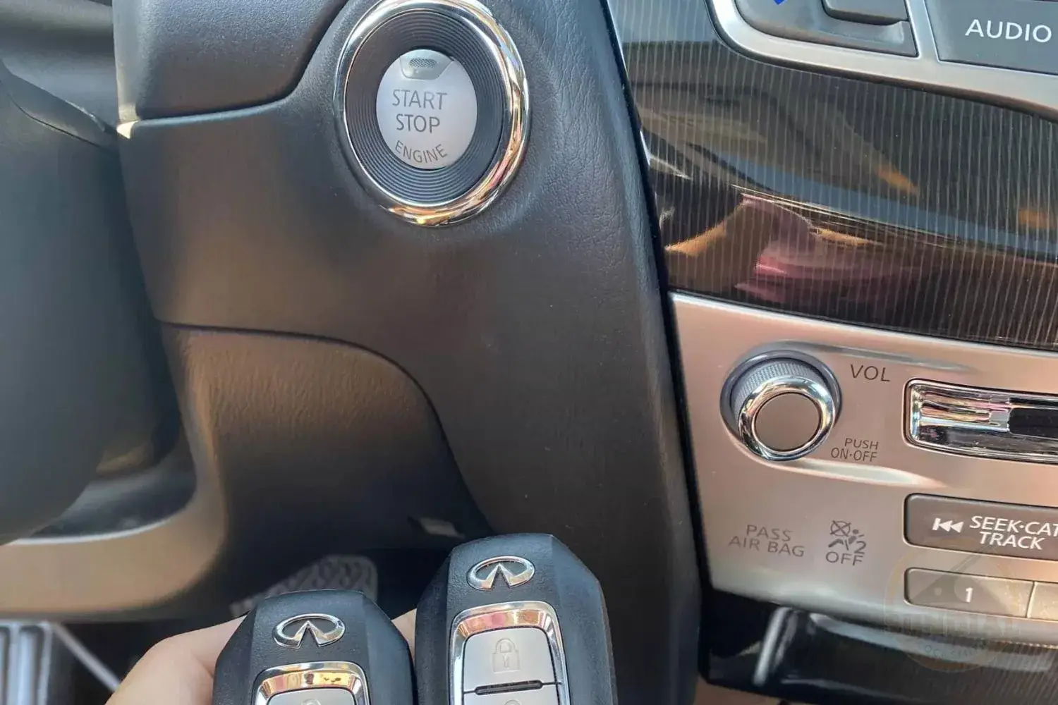 A close-up of two key fobs held near the start/stop engine button in a car's interior, with audio controls in the background