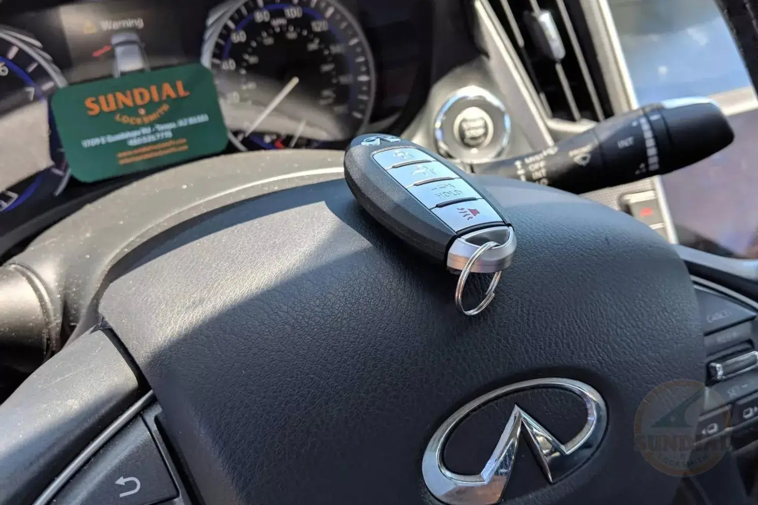 A car key fob placed on the leather steering wheel of a luxury car with the vehicle's dashboard and a locksmith's business card visible in the background.