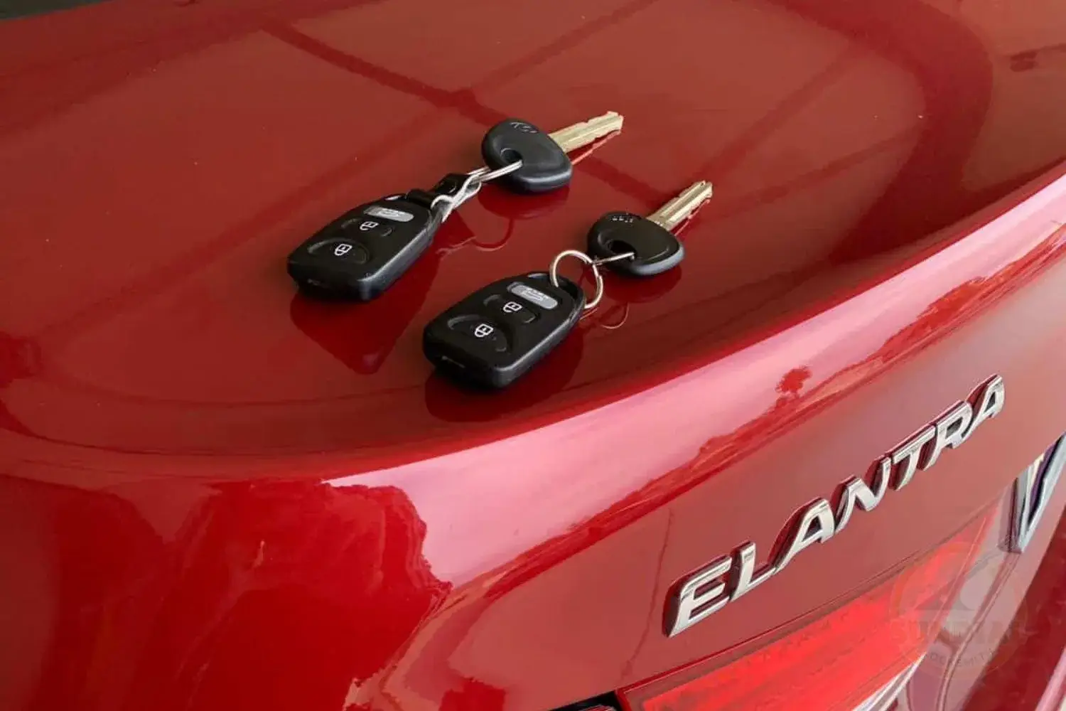 Two Hyundai car key fobs with attached keys resting on the trunk lid of a red Hyundai Elantra.