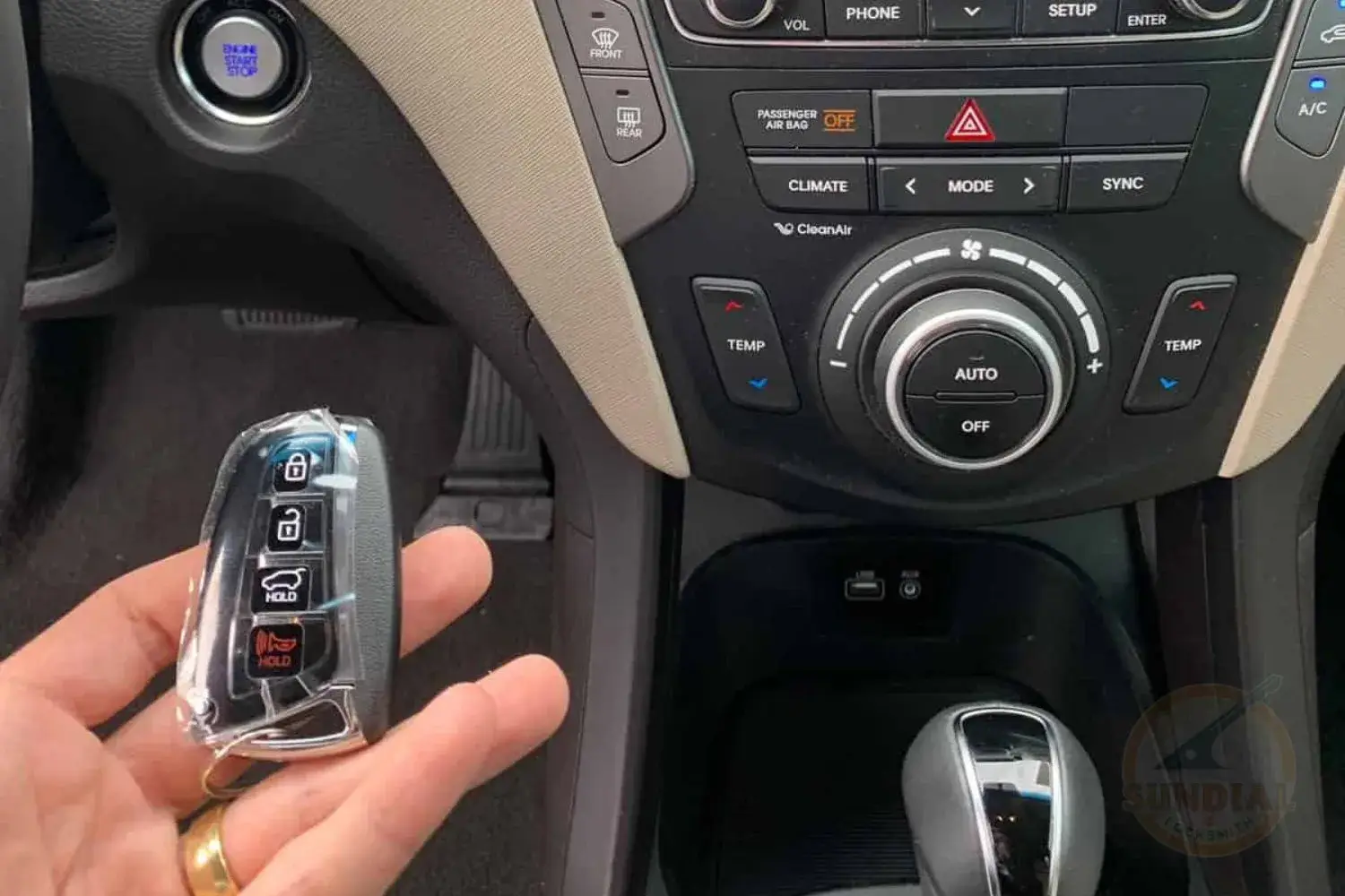 A person's hand holding a new Hyundai key fob, still in its protective cover, inside the vehicle near the start button and climate control panel.