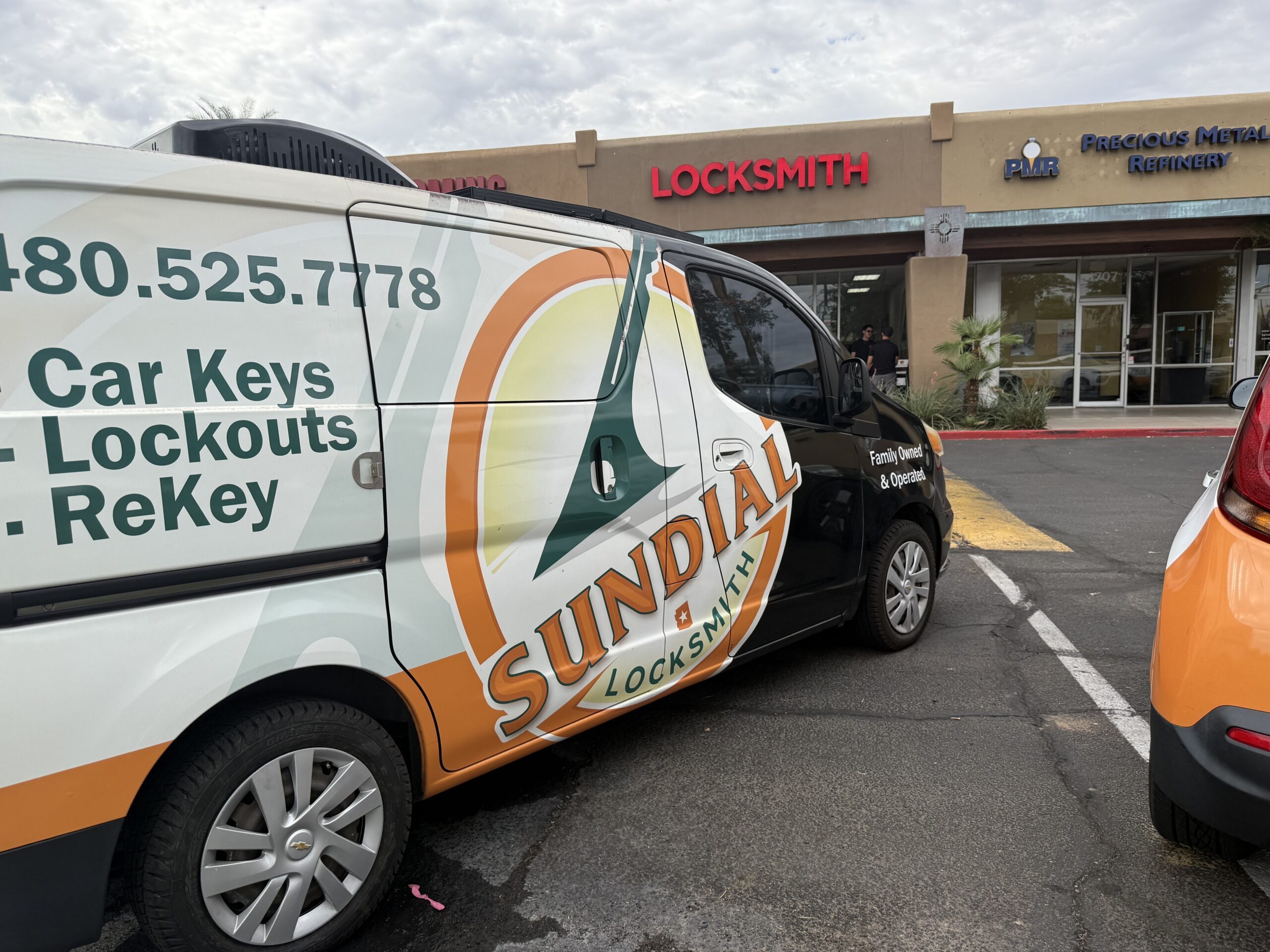 A Sundial Locksmith van is parked in front of a strip mall. The van displays services like car keys, lockouts, and rekeying. The Locksmith shop sign is visible on a nearby storefront.