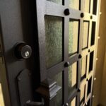 A close-up view of a partially open black metal door with a grid pattern and textured glass panels. The door handle and lock are visible, with warm light illuminating the scene from inside.