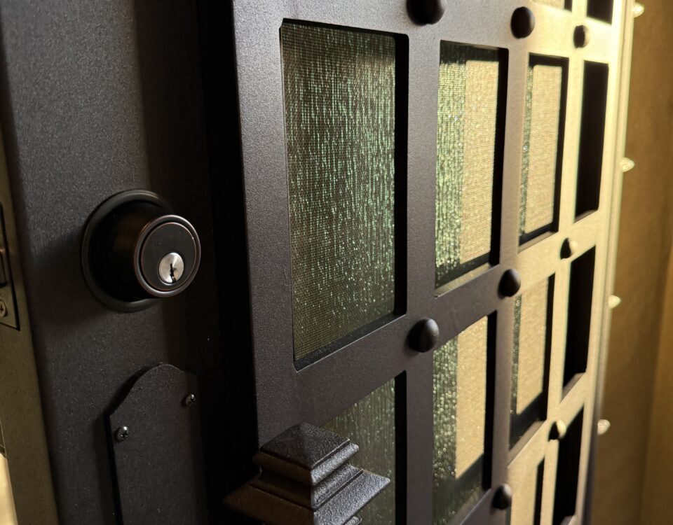 A close-up view of a partially open black metal door with a grid pattern and textured glass panels. The door handle and lock are visible, with warm light illuminating the scene from inside.