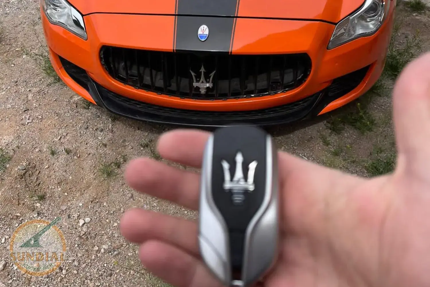 A hand holding a Maserati car key with an orange Maserati vehicle in the background.