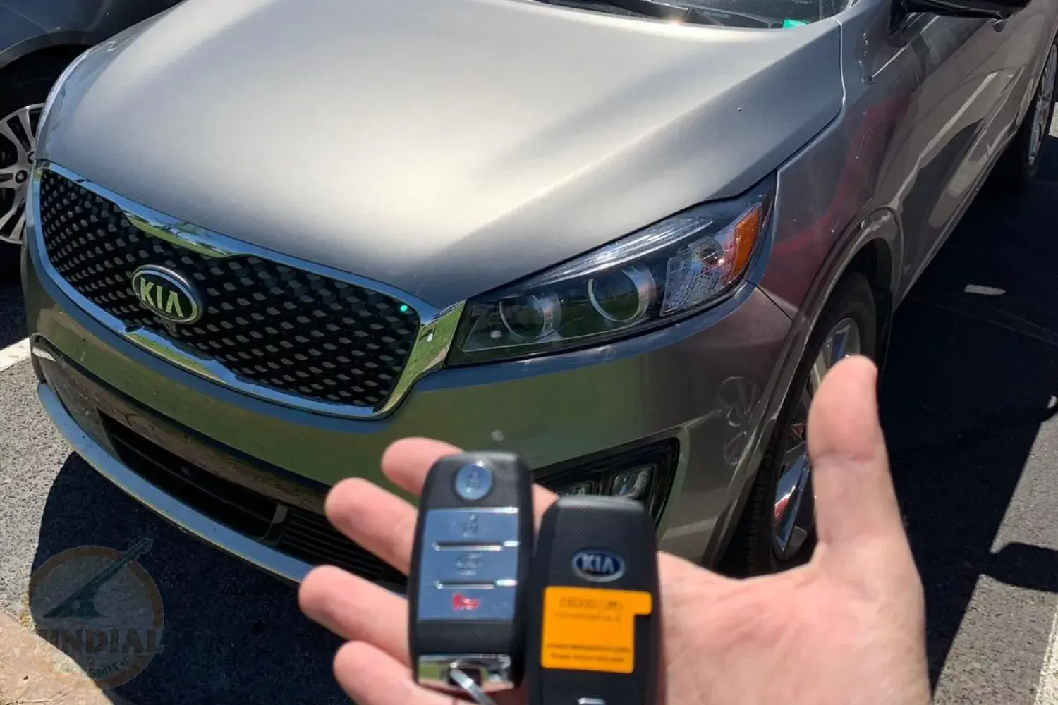 A hand holding two Kia key fobs in front of a metallic gray Kia car.